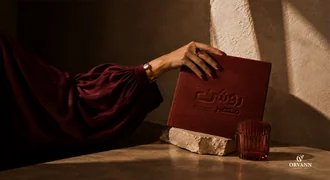 A hand holding a burgundy book embossed with the Roshan Masr logo.